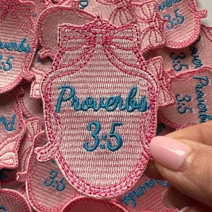 May include: A pink fabric patch with blue embroidery that reads "Proverbs 3:5". The patch is shaped like a heart with a bow at the top.