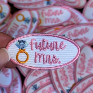 May include: A close-up of a pink and white oval-shaped embroidered patch with the text "Future Mrs." in white cursive lettering. The patch features a gold ring with a blue diamond.