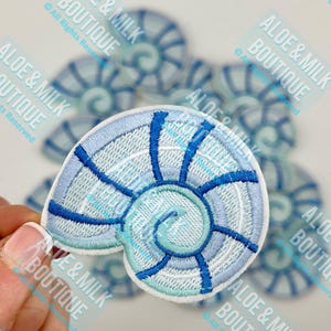 May include: A set of five blue and white embroidered iron-on patches shaped like seashells. The patches are all the same design, with a blue and white spiral pattern.