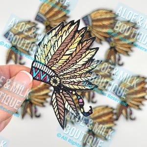 May include: Embroidered patch in the shape of a Native American headdress. The patch features a detailed design with brown, yellow, teal, and red colors. The patch is held in a hand, with other patches in the background.