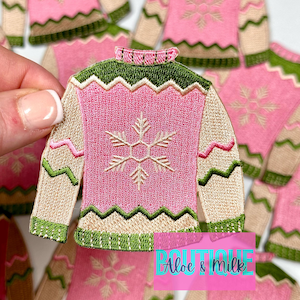 May include: A pink and green embroidered iron-on patch of a Christmas sweater with a snowflake design. The patch is for crafting and decorating.