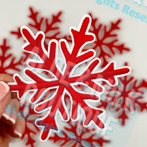 May include: Red and white embroidered snowflake patch. The snowflake has a detailed design with a red center and white outline. Several other snowflakes are visible in the background, creating a festive winter theme.