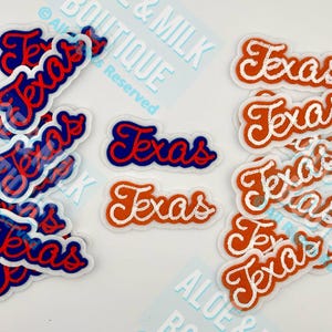 May include: A collection of embroidered patches with the word "Texas" in a script font. The patches are in red, white, and blue, with a clear border. Some patches are stacked, and others are scattered on a white surface.