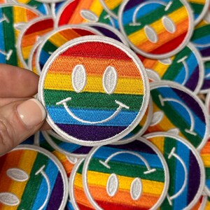 May include: A close-up of a rainbow smiley face patch. The patch is made of embroidered fabric and has a white outline. The rainbow colors are red, orange, yellow, green, blue, indigo, and violet.