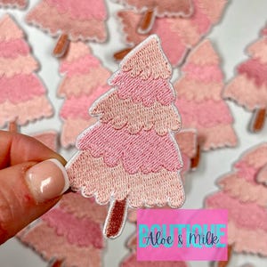 May include: A set of pink and white felt Christmas tree patches. The trees are layered and have a brown felt trunk. The patches are perfect for adding a festive touch to clothing, bags, or other crafts.