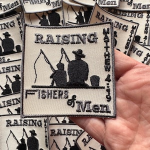 May include: A white embroidered patch with the text "Raising Fishers of Men Matthew 4:19" and a black silhouette of three people fishing. The patch has a gray border.