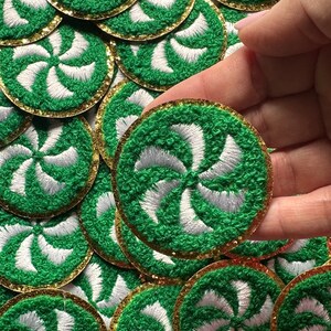 May include: Embroidered green and white circular patches with a gold glitter border. The patches feature a swirling design in white and are densely stitched. A hand holds one patch, with many more visible in the background.