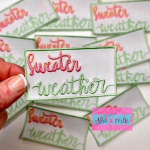 May include: A white iron-on patch with green and pink embroidered text that reads "sweater weather".