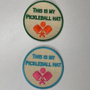 May include: Two round embroidered patches with beige fabric and colorful embroidery. The top patch has a green border and orange paddles. The bottom patch has a blue border and pink paddles. Both patches say "This is my pickleball hat".