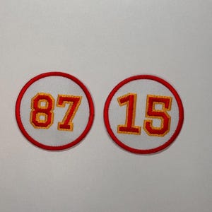 May include: Two white, round, embroidered patches with red borders. The patches feature the numbers "87" and "15" in yellow thread.