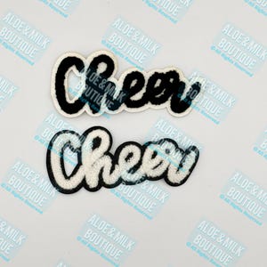 May include: Two black and white felt patches with the word "Cheer" in a cursive font. The patches are designed to be sewn or ironed onto clothing or other items.
