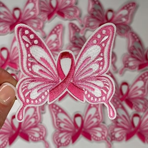 May include: Pink butterfly-shaped iron-on patches with a pink ribbon in the center. The patches are designed to show support for breast cancer awareness.