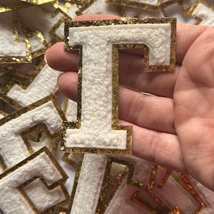 May include: White felt letter I with a gold glitter outline. The letter is held in a hand.