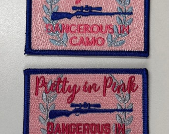 Puede incluir: Dos parches bordados de color rosa y azul con el texto "Pretty in Pink Dangerous in Camo" y un rifle con mira telescópica.