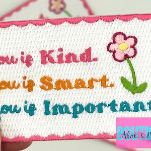 May include: Embroidered patch with the words "You is Kind, You is Smart, You is Important" in pink, orange, and teal. A flower with pink petals and a green stem is on the right. The patch has a pink scalloped border.