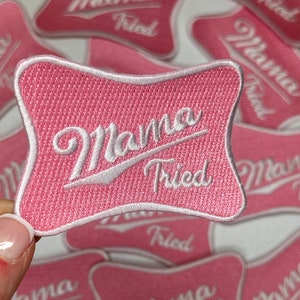 May include: Pink embroidered patch with the words "Mama Tried" in white script. The patch has a unique shape with a white border. Several similar patches are visible in the background.
