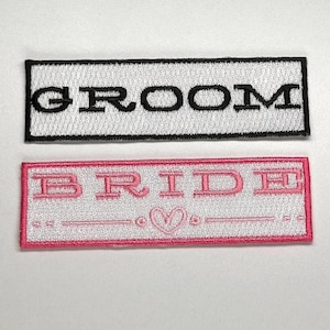 May include: Two white fabric patches with black and pink embroidery. The top patch says "GROOM" in black lettering. The bottom patch says "BRIDE" in pink lettering with a heart symbol between two lines.