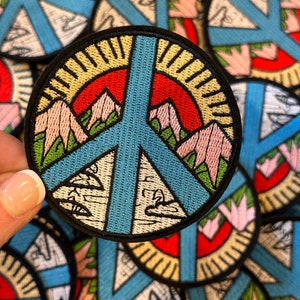 May include: A close-up of a peace sign embroidered patch with a mountain scene and a sun. The patch is blue, green, pink, red, and yellow. The patch is surrounded by other similar patches.