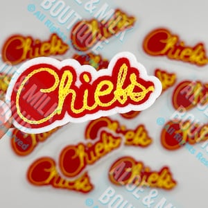 May include: A collection of red and yellow "Chiefs" patches. The word "Chiefs" is embroidered in yellow thread on a red background, outlined in white. Several patches are scattered across the image, with a hand holding one in the foreground.