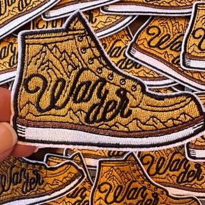 May include: A yellow and brown embroidered patch of a hiking boot with the text "Wander" in a stylized font. The boot is surrounded by a white border.
