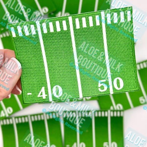 May include: Green embroidered patch depicting a football field with white yard lines and numbers. The patch shows the 40 and 50-yard lines. The patch is rectangular and has a stitched border. The background is a light color.