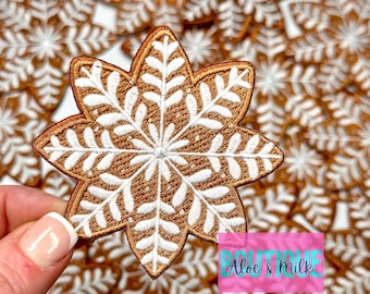 May include: A close-up of a pile of brown and white snowflake-shaped iron-on patches. The patches are embroidered with a white snowflake design on a brown background. The text "Aloe & Milk" is visible in the lower right corner of the image.