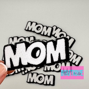 May include: A close-up of multiple white felt patches with the word "MOM" in black outlined letters. The patches are arranged in a pile, with the largest patch in the center.