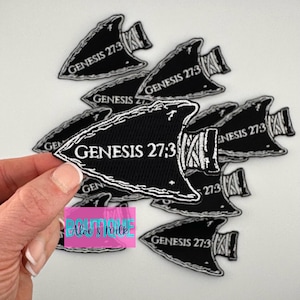May include: Black arrowhead patches with white text that reads "GENESIS 27:3". The patches have a white border and a woven texture. The image includes a hand holding one patch, with others scattered in the background.