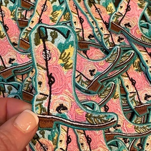 May include: A stack of pink and turquoise embroidered iron-on patches in the shape of a cowboy boot with floral designs.