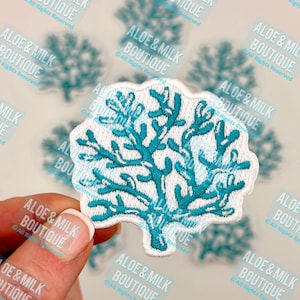 May include: A white embroidered patch with a turquoise blue coral design. The patch is held in a hand with a white manicure.