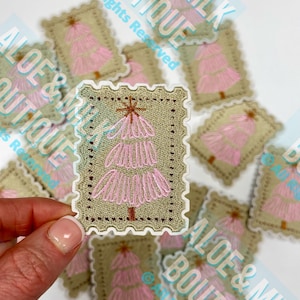 May include: A collection of embroidered patches featuring a pink Christmas tree design. The patches are rectangular with a postage stamp-like border and a tan background. The tree has a star on top and a brown trunk. The text "ALOE & MILK BOUTIQUE" is visible.