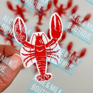 May include: A red and white embroidered iron-on patch of a lobster. The lobster is facing right and has a white background. The patch is surrounded by other lobster patches.