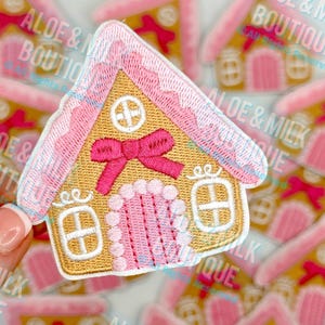 May include: Embroidered patch featuring a gingerbread house design. The house has a tan base with pink roof and door, and white accents. A pink bow adorns the front. The patch is surrounded by other similar patches.