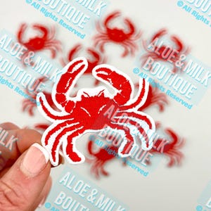 May include: Red embroidered iron-on patch of a crab with white outlines. The patch is on a white background. The text "ALOE & MILK BOUTIQUE" is visible in the background.