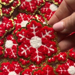 May include: Red and white snowflake-shaped appliqués with gold glitter edging. The appliqués are made of a textured material and are arranged in a pile. One is being held up by a hand.