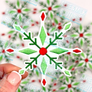 May include: Embroidered snowflake patch with a white background, featuring green diamond shapes, red accents, and a red center. The patch has a festive, holiday theme. The text "ALOE & MILK BOUTIQUE" is visible.