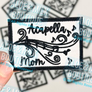 May include: A rectangular patch with a white background and black embroidered text and design. The text reads "Acapella Mom" with musical notes and a treble clef. The patch is held by a hand.