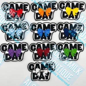 May include: A collection of fabric patches with the words "GAME DAY" in black block letters, each featuring a colorful bow. The bows come in various colors, including red, orange, yellow, blue, purple, green, and white.