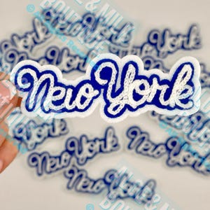 May include: A white and blue embroidered patch with the words "New York" in a cursive font. The patch has a blue border and white stitching. Several similar patches are scattered in the background.