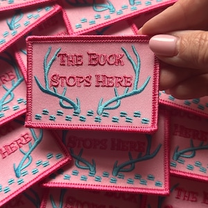 May include: A pink fabric patch with a blue embroidered design of a buck's antlers and the text "The Buck Stops Here".