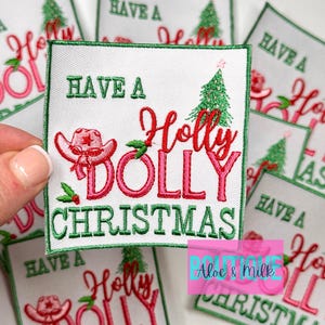 May include: A white fabric patch with green embroidered trim. The patch features a pink and red embroidered design of a cowboy hat, a Christmas tree, and the text "Have a Holly Dolly Christmas".