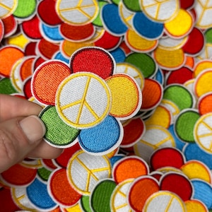 May include: A colorful flower-shaped patch with a peace sign in the center. The flower is made of six petals in red, orange, yellow, green, blue, and white.