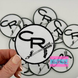 May include: A set of white, round iron-on patches with black embroidery. The patches feature the letters "CR" with an arrow pointing to the right. The text "Aloe & Milk Boutique" is visible in the bottom right corner of the image.