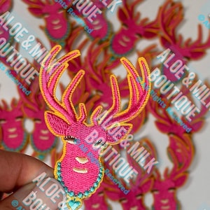 May include: A close-up of several pink and yellow embroidered iron-on patches featuring a cartoon deer with eyelashes and a turquoise beaded necklace. The patches are labeled "Aloe & Milk Boutique" with the text "All Rights Reserved".