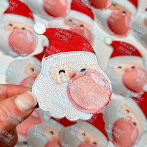 May include: A red and white embroidered iron-on patch of Santa Claus blowing a pink bubble. The patch is surrounded by other Santa Claus patches.