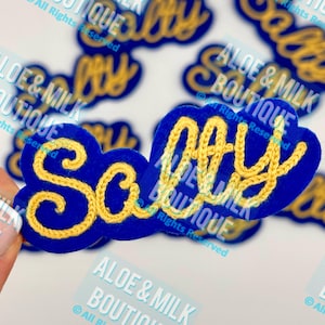 May include: A close-up of a blue felt patch with the word "Sally" embroidered in yellow thread. The patch is surrounded by other patches with the same design but different names.