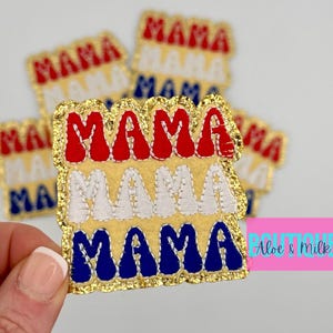 May include: Embroidered patches with the word "MAMA" in red, white, and blue, layered on a gold background. The patches have a gold glitter border. Several patches are visible, with one held by a hand.