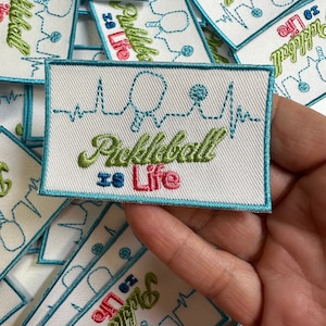 May include: A white patch with blue trim features an embroidered green and pink design of a heartbeat with a paddle and ball. The text "Pickleball is Life" is embroidered in green and pink.