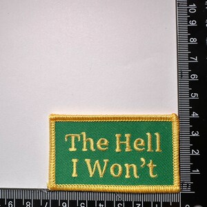 May include: A green embroidered patch with a gold border. The patch says "The Hell I Won't" in gold thread.