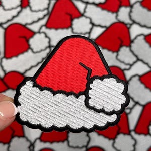May include: A red and white embroidered Santa hat patch. The patch is made of felt and has a black outline.
